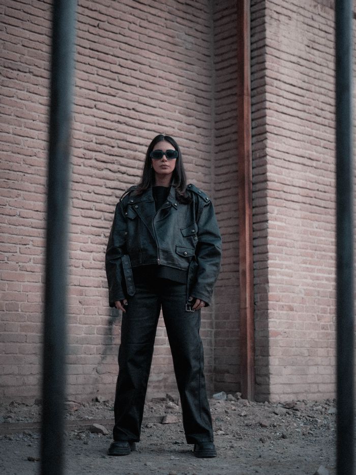 Stylish woman in black leather jacket and sunglasses posing against a rustic brick wall.