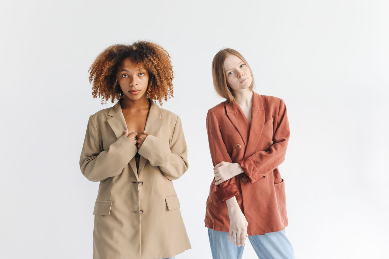services-01 Two women modeling fashionable blazers in a studio setting with a white backdrop.