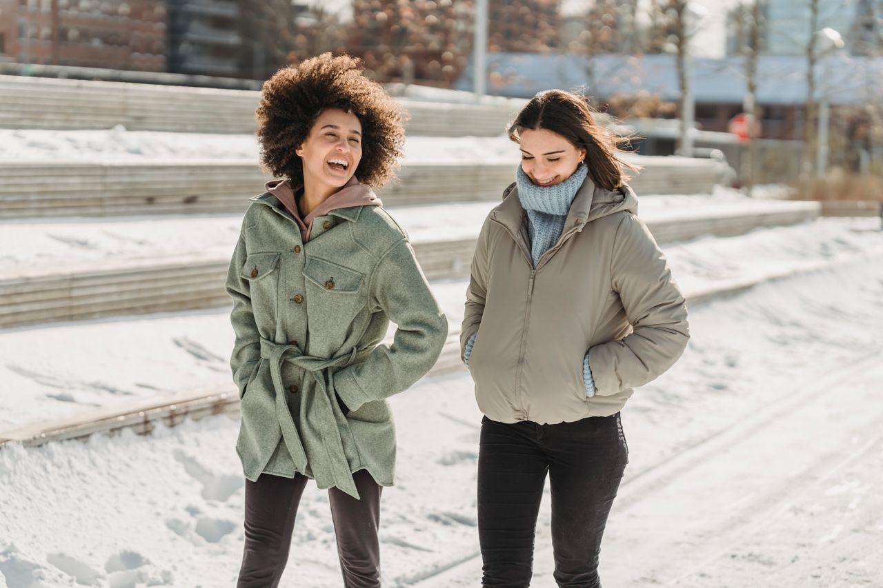 services-04 Expressive young multiracial female friends wearing warm clothes chatting and walking together with hands in pockets on snowy city street on freezing winter day